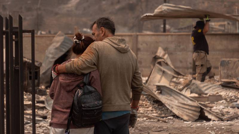 Una pareja se abraza mientras un hombre carga escombros de una casa incendiada.
